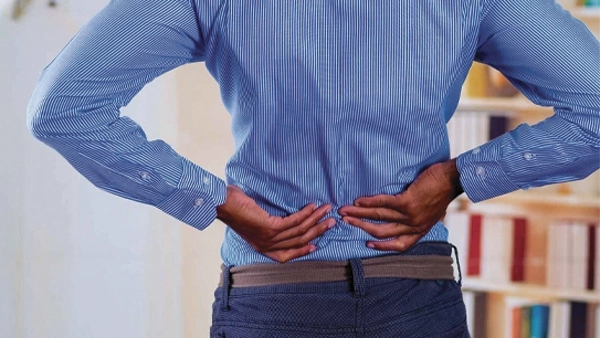 Chiropractic Timonium MD Back pain HP person in formal clothing holding their lower back, illustrating discomfort or back pain in an indoor setting.